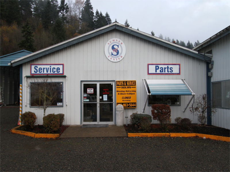 Outside of Sumner RV's service department, with a yellow sign next to the front door and trees in …